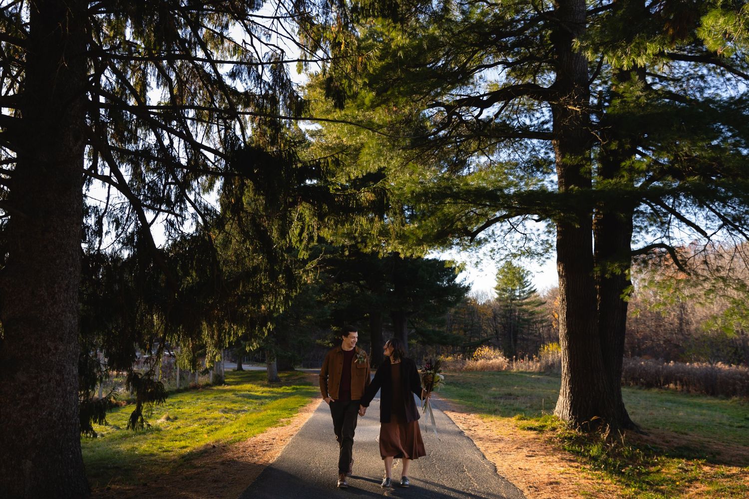 Two people walk together on a sunlit path through a scenic park with tall pine trees on a beautiful autumn day.