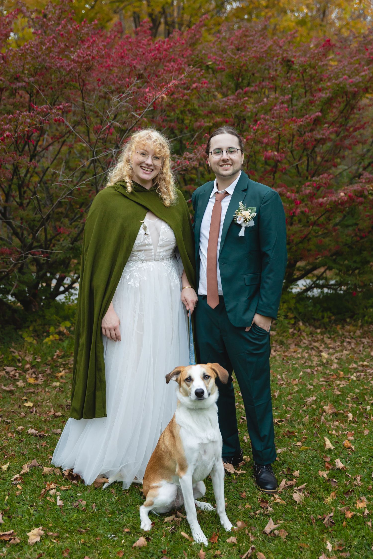 Couple in wedding attire with green cape posing with their dog against autumn foliage.