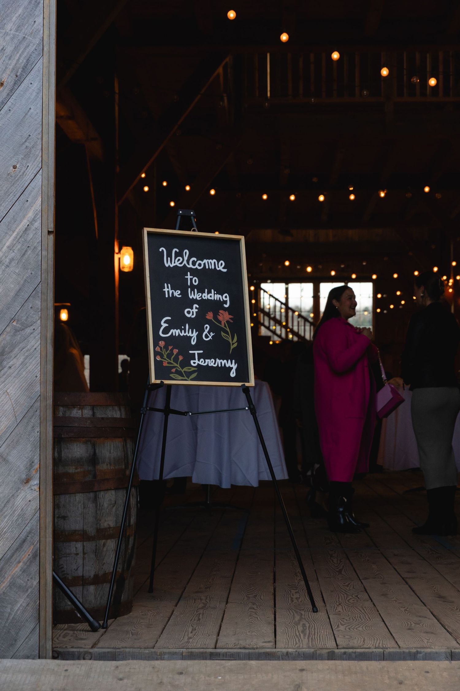 A chalkboard wedding welcome sign stands at the entrance of a dimly lit rustic venue with string lights overhead.