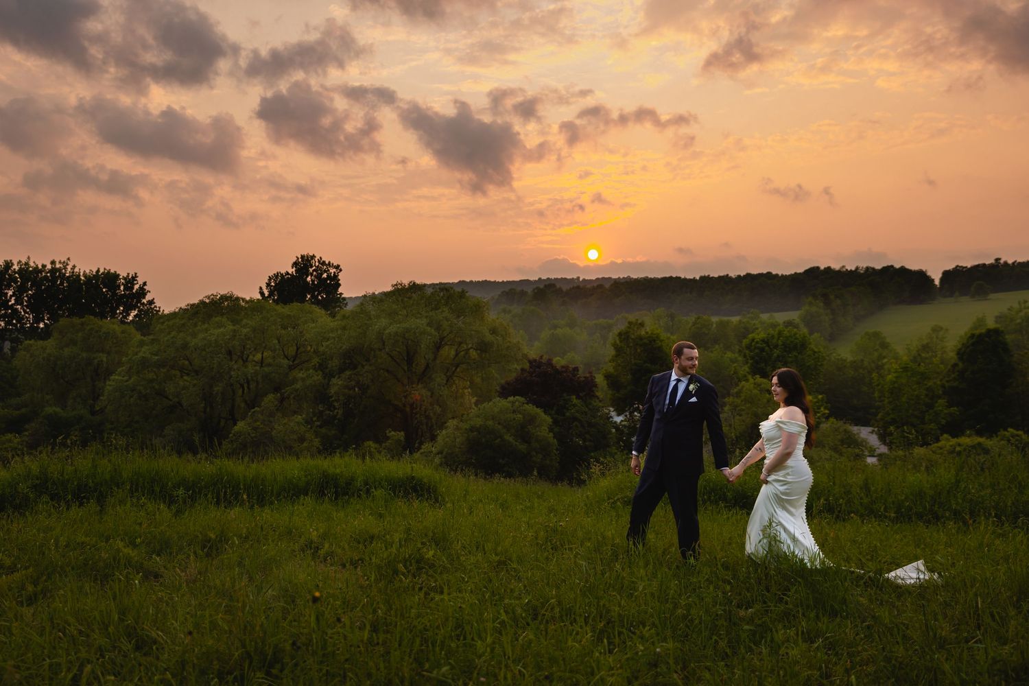 A couple in wedding attire walks through a scenic meadow at sunset with rolling hills in the background.