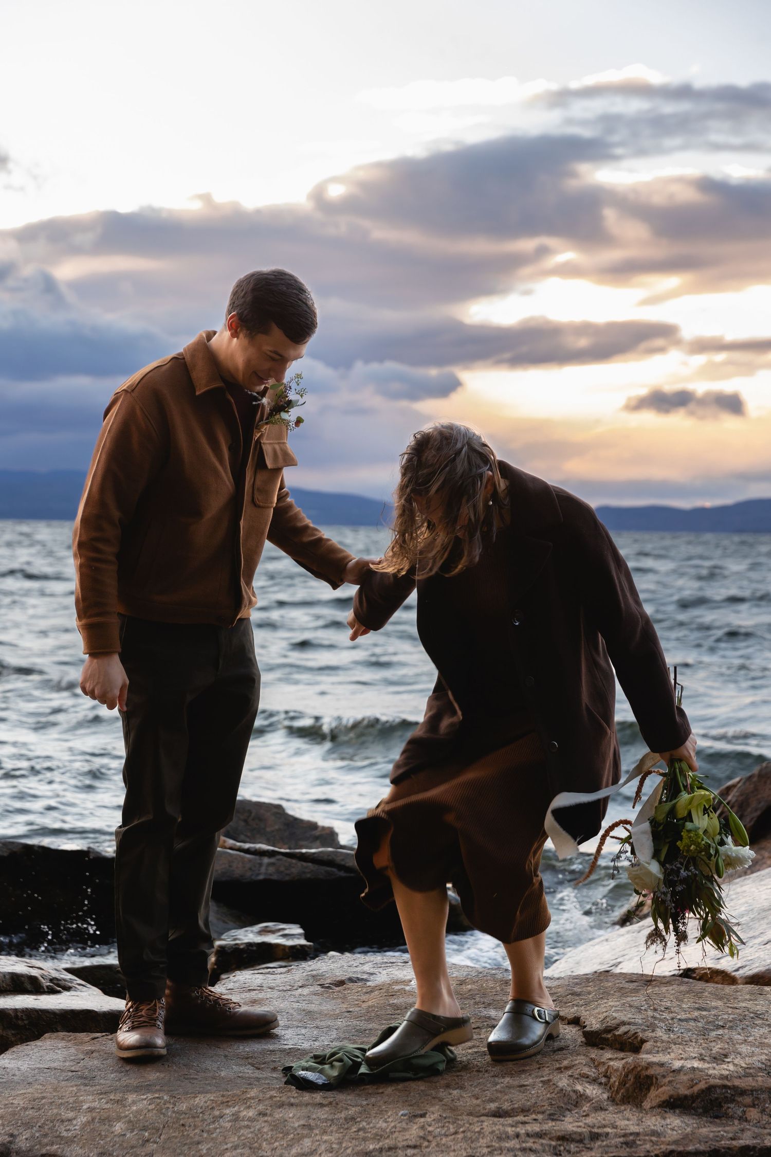 Two people walking carefully on rocky coastal shore during sunset, with choppy waters and dramatic clouds in background.