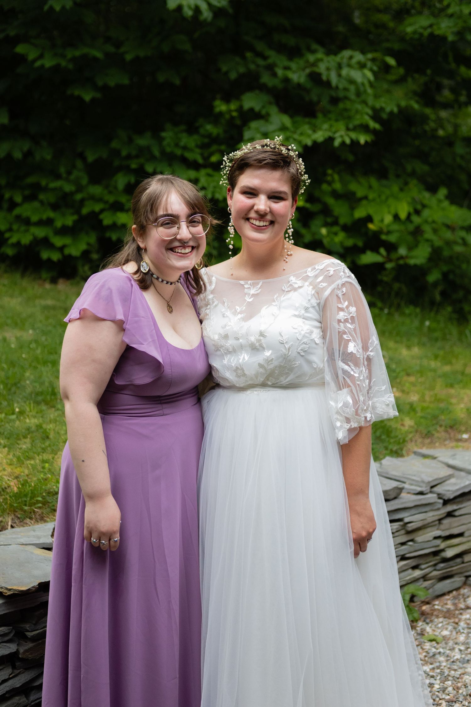 Wedding portrait with bride in lace gown and attendant in purple dress against garden backdrop.