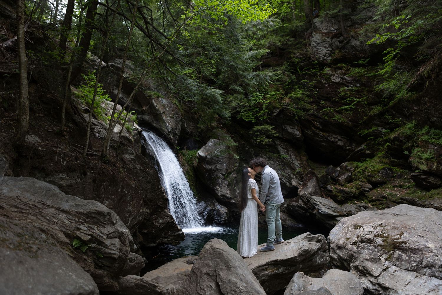 A couple stands on rocky terrain before a cascading waterfall in a dense forest setting.