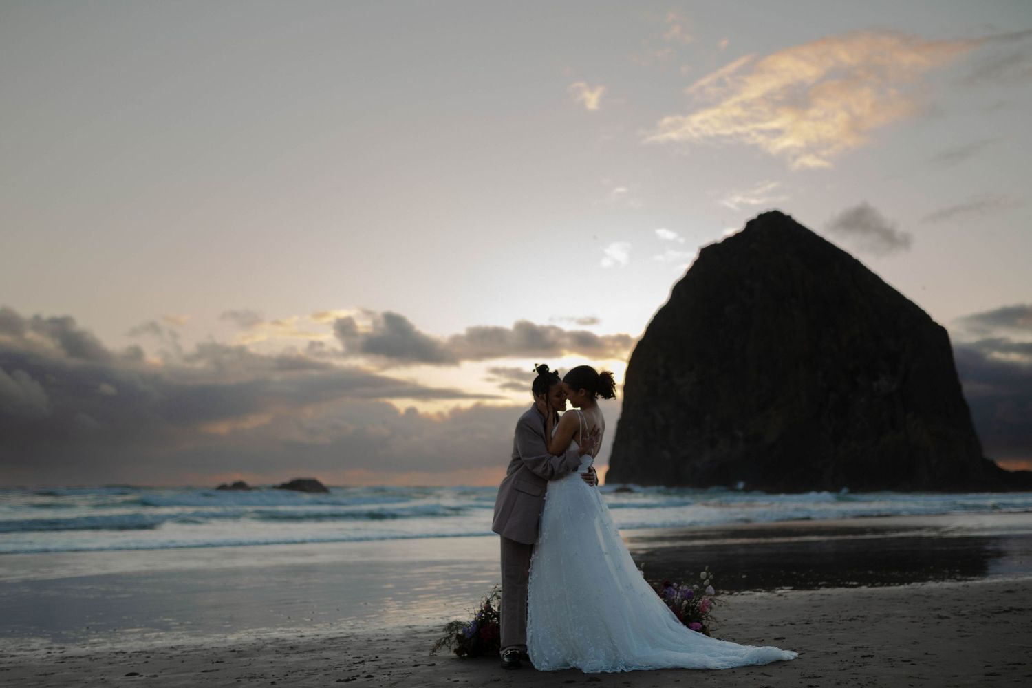 A couple shares an intimate sunset moment on a beach with the iconic Haystack Rock at Cannon Beach in the background.