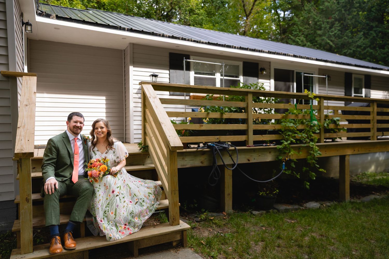 A couple sits on wooden deck stairs outside a ranch-style house surrounded by greenery.