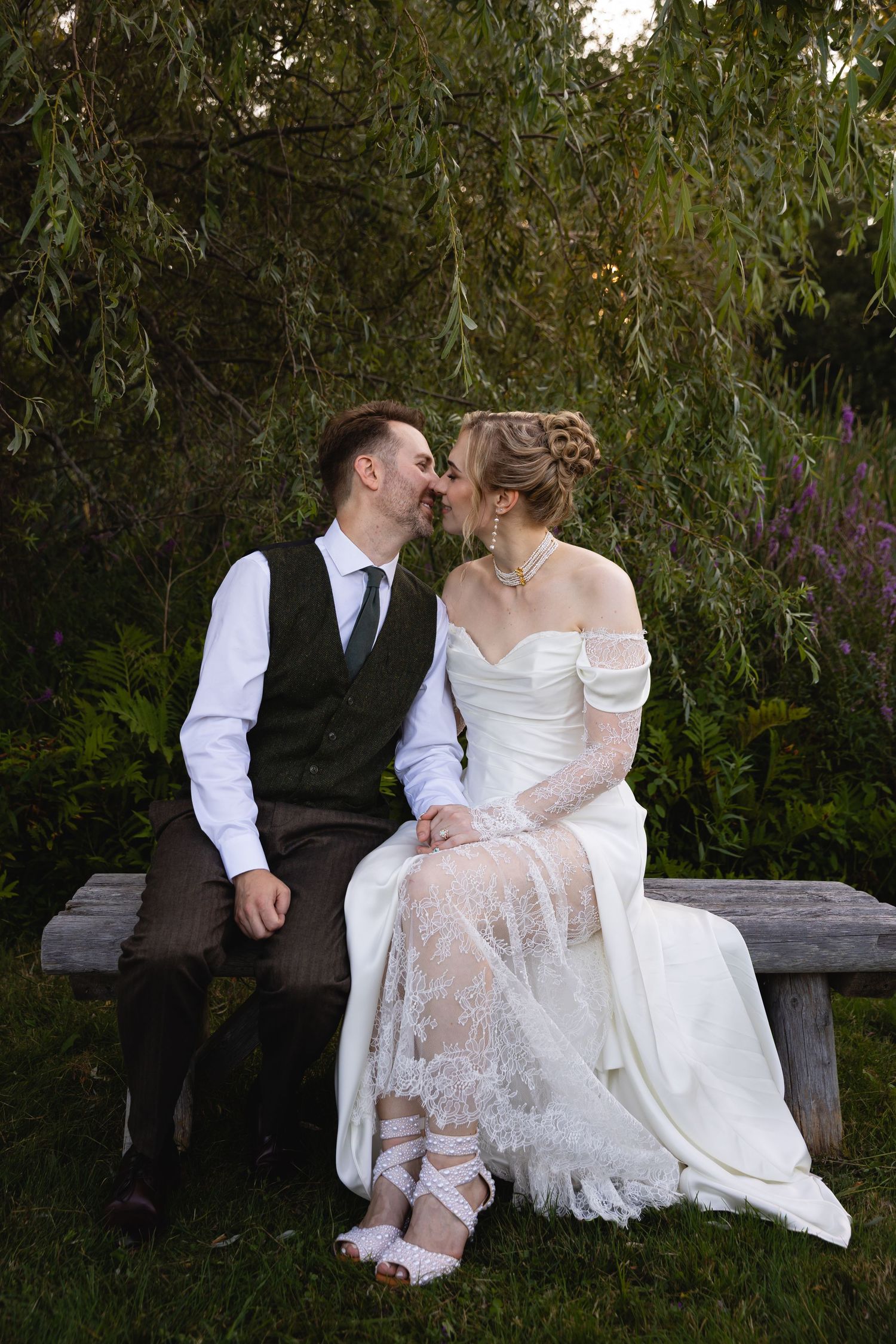 A couple in wedding attire share a romantic moment on a wooden bench under a willow tree in a garden setting.