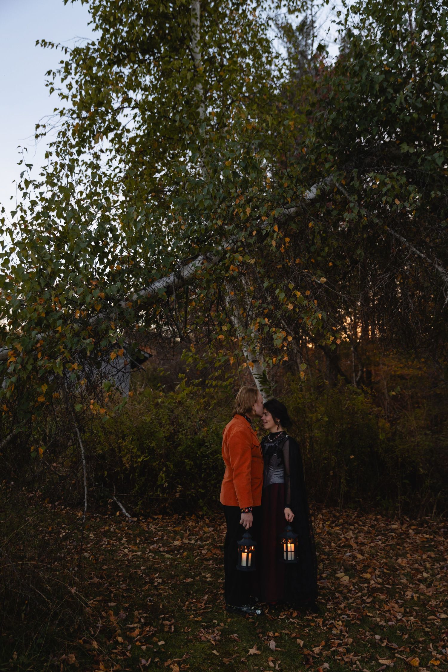 Two people stand together in a wooded area covered with fallen autumn leaves at dusk.