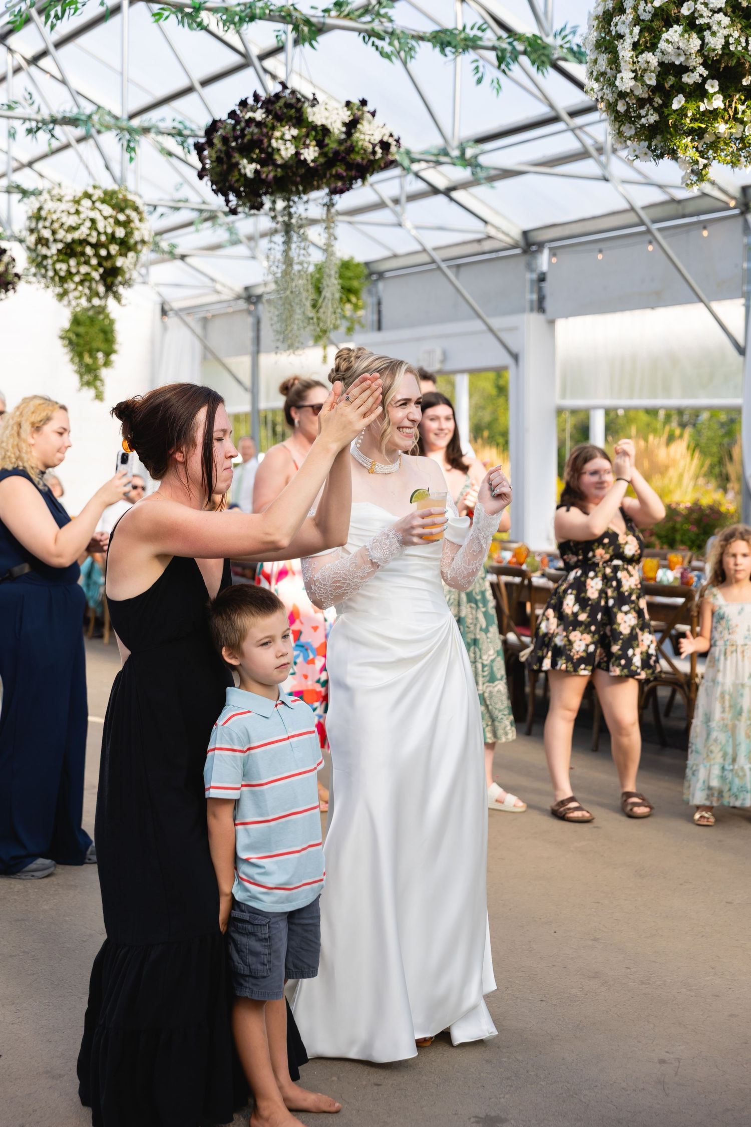 Wedding guests dance and celebrate together in a bright greenhouse reception venue during a summer celebration.