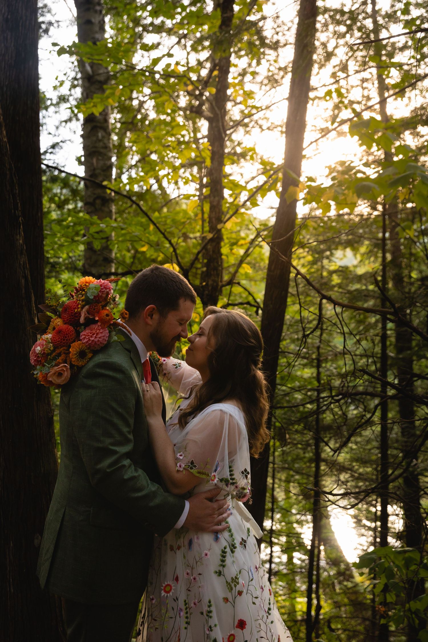 Romantic couple shares an intimate moment in a sunlit forest wearing floral dress and suit.