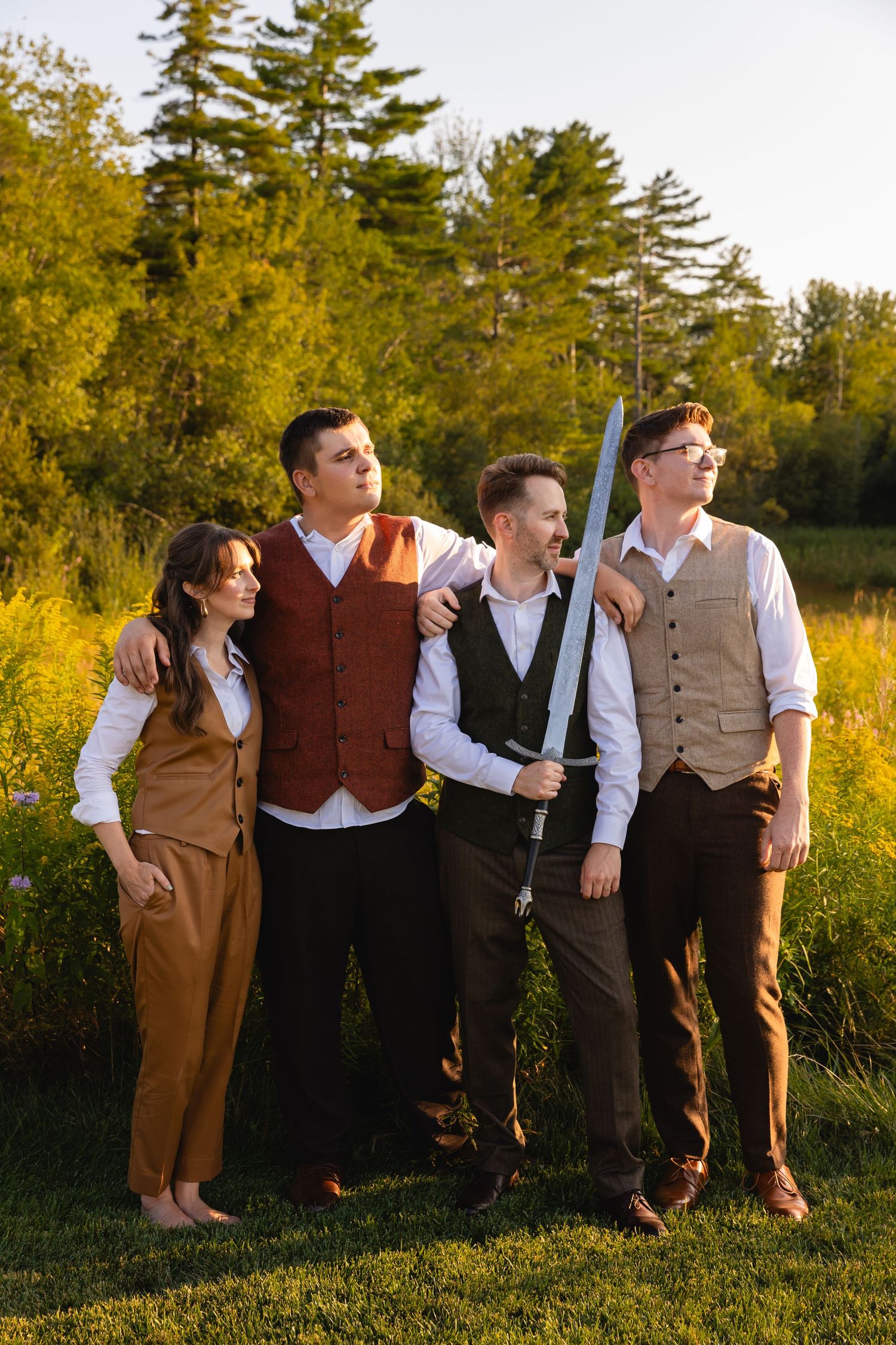 Wedding party of four posing outdoors at golden hour, one holding a decorative sword.