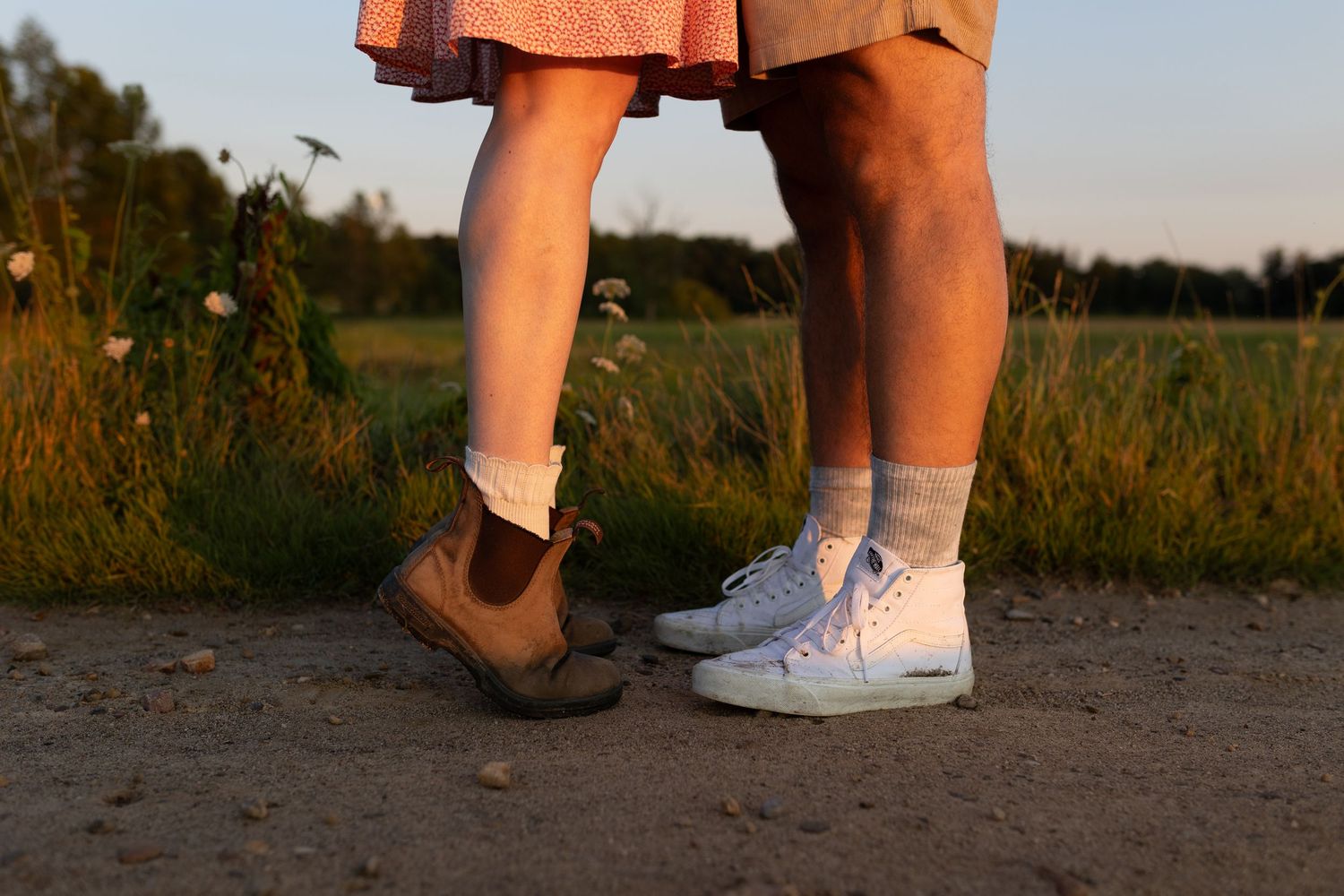 Close-up of a couple's feet on a dirt path, one wearing brown boots and the other white sneakers at sunset.