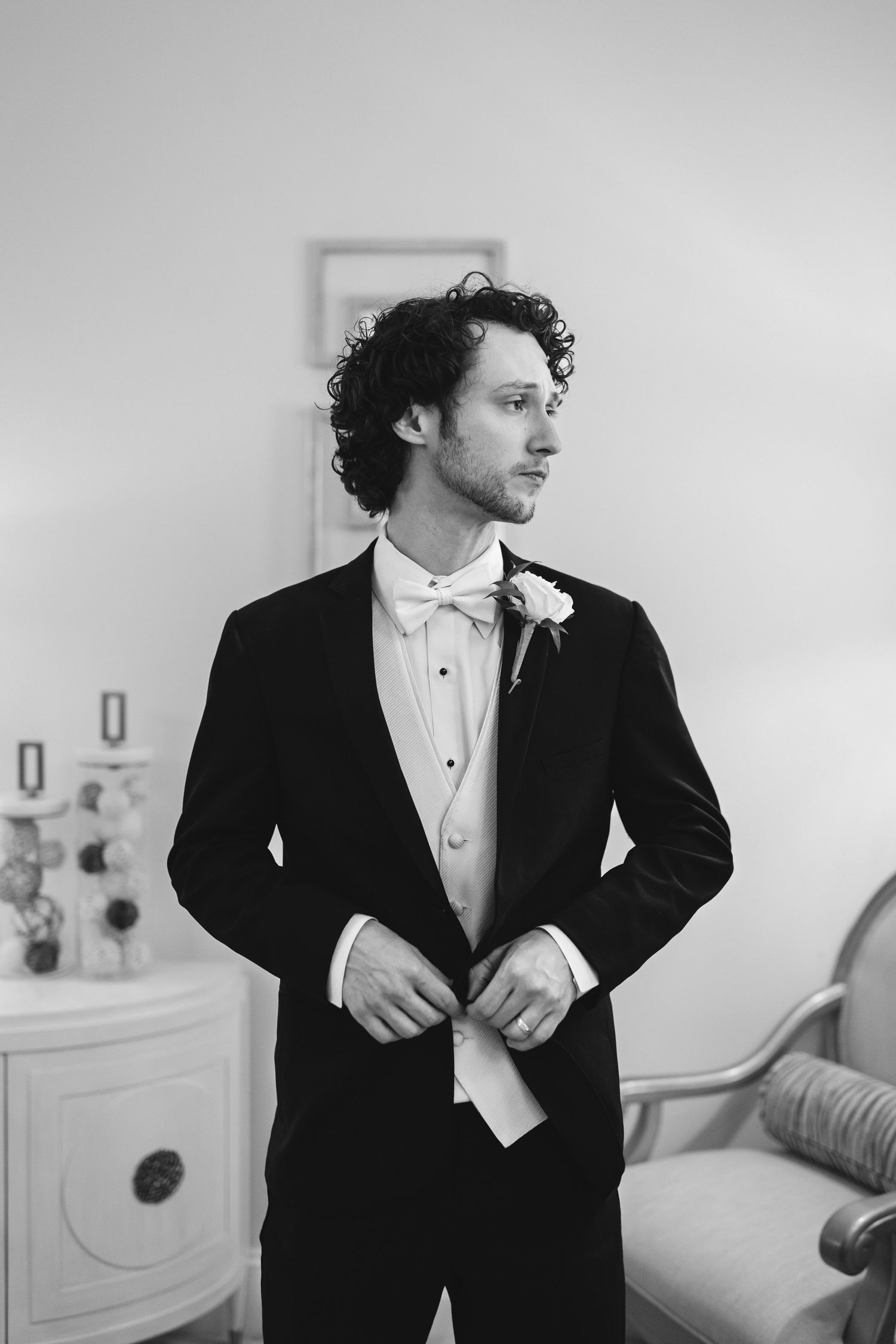 A groom in a black tuxedo and bow tie with curly hair adjusts his cuffs in a black and white portrait.