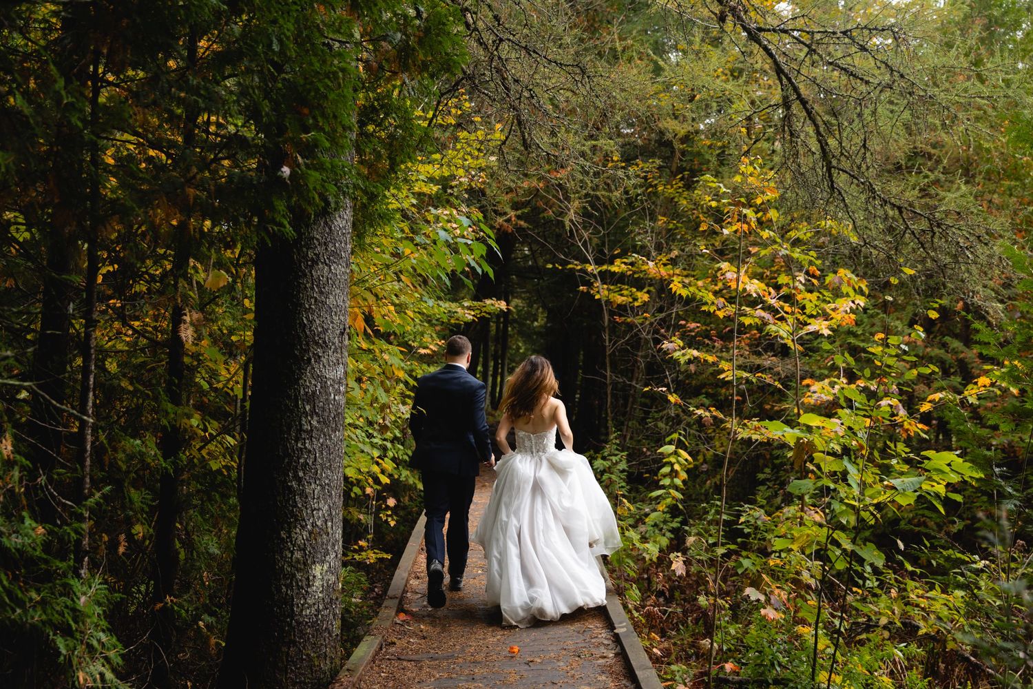 A bride in a flowing white gown walks along a forested trail surrounded by autumn foliage.