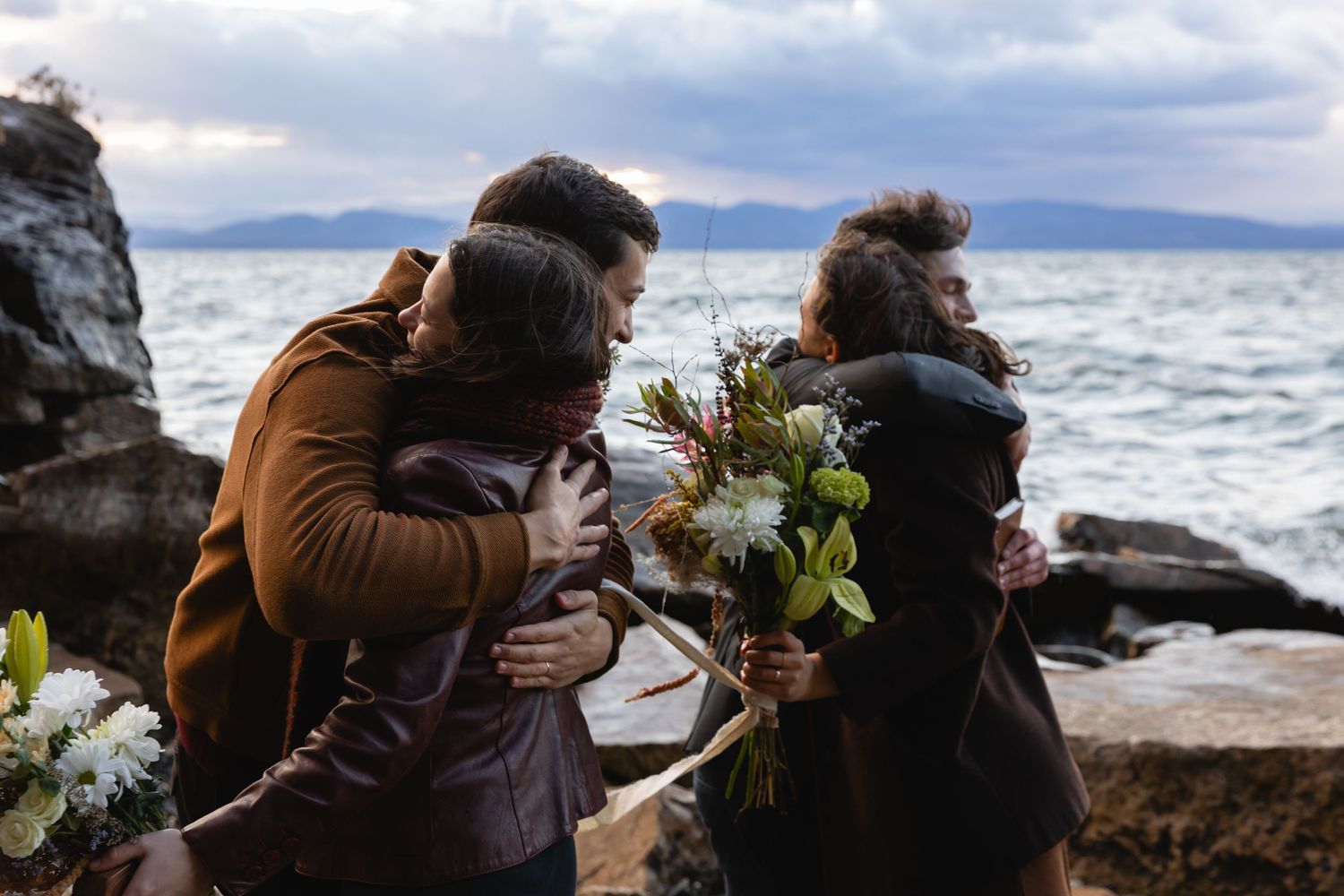 A couple shares a tender moment on rocky coastal cliffs while holding white floral bouquets by the ocean.