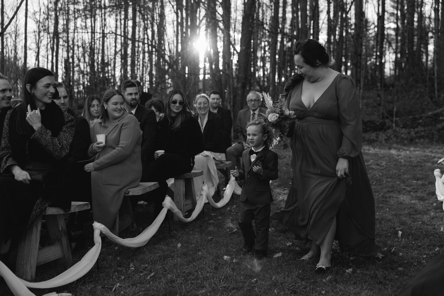Vintage black and white photograph showing people gathered outdoors in a forest clearing with sun peeking through trees.
