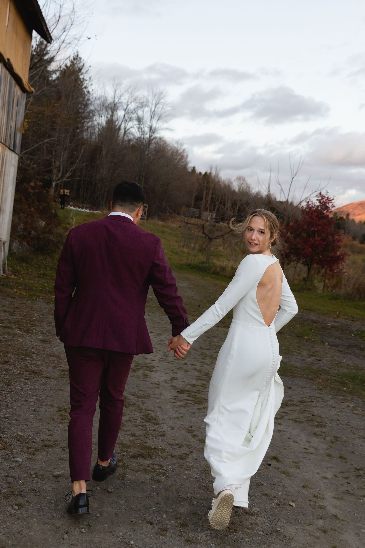A couple holding hands and walking on a dirt path, with the groom in burgundy suit and bride in white gown.