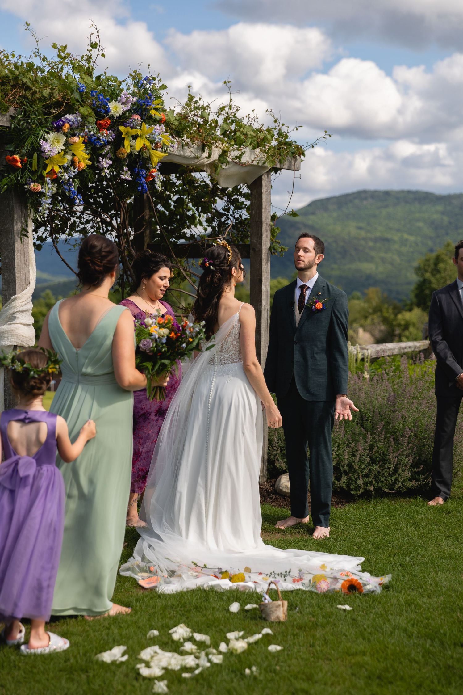 A wedding ceremony takes place under a flower-adorned arbor with mountain views.