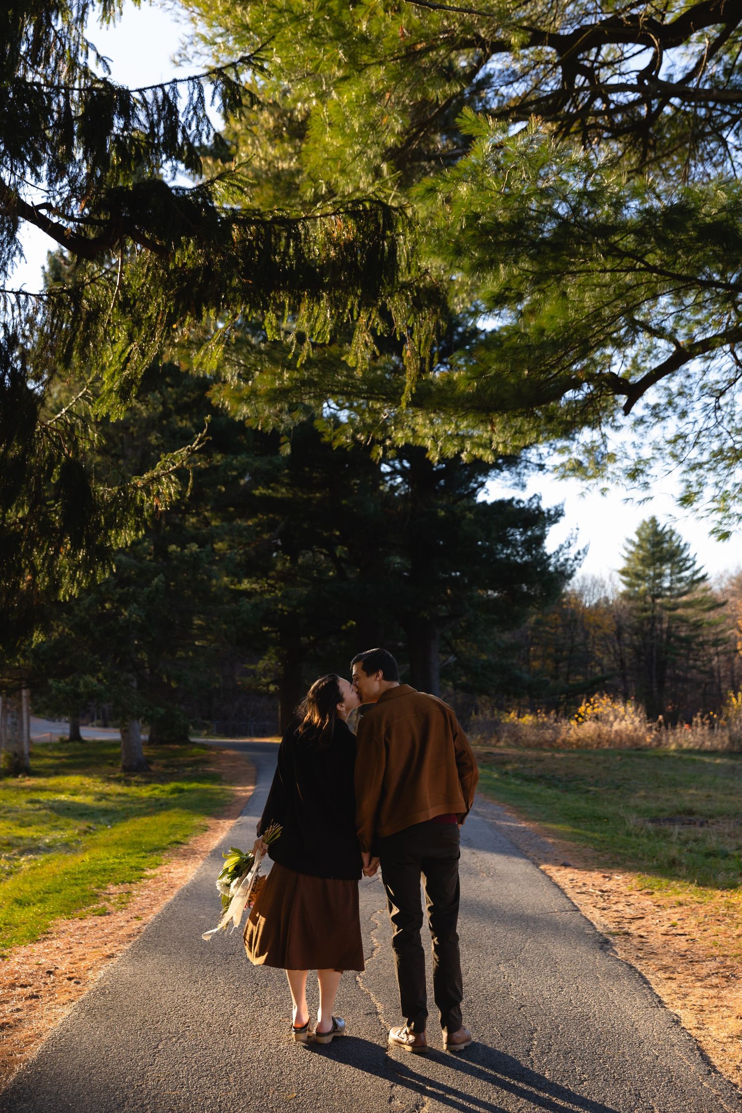 Two people walk together down a sunlit path surrounded by towering evergreen trees on an autumn afternoon.