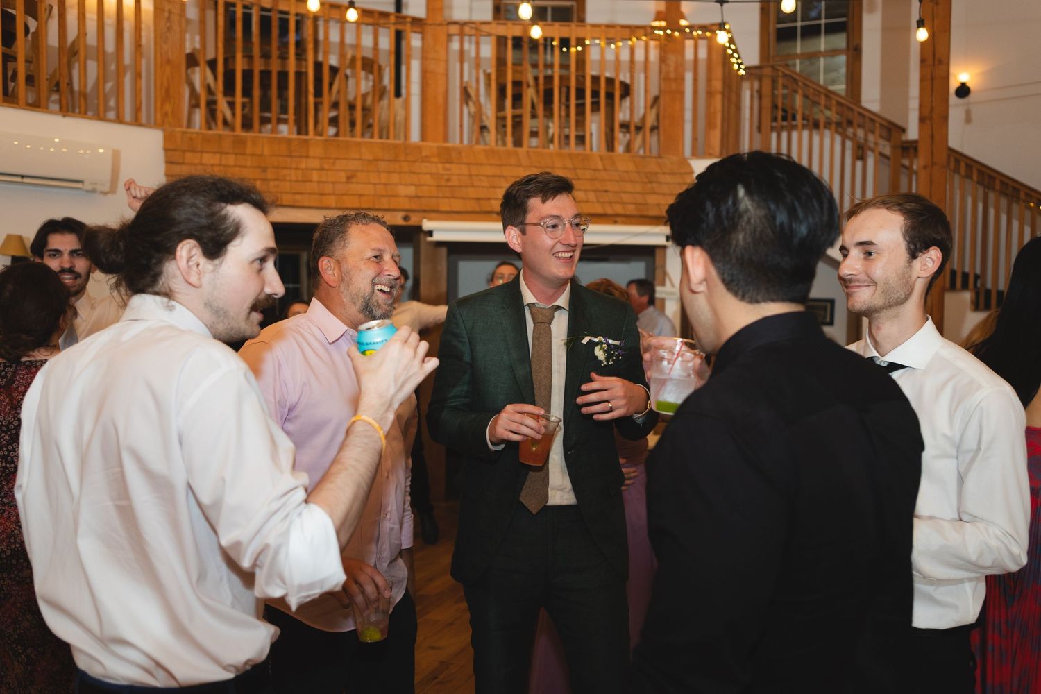 A group of guests enjoying drinks and conversation at an indoor celebration with wooden stairs in the background.