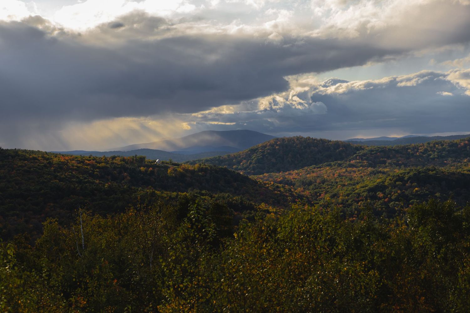 Sunlight breaks through dark clouds over a mountainous landscape with fall foliage.