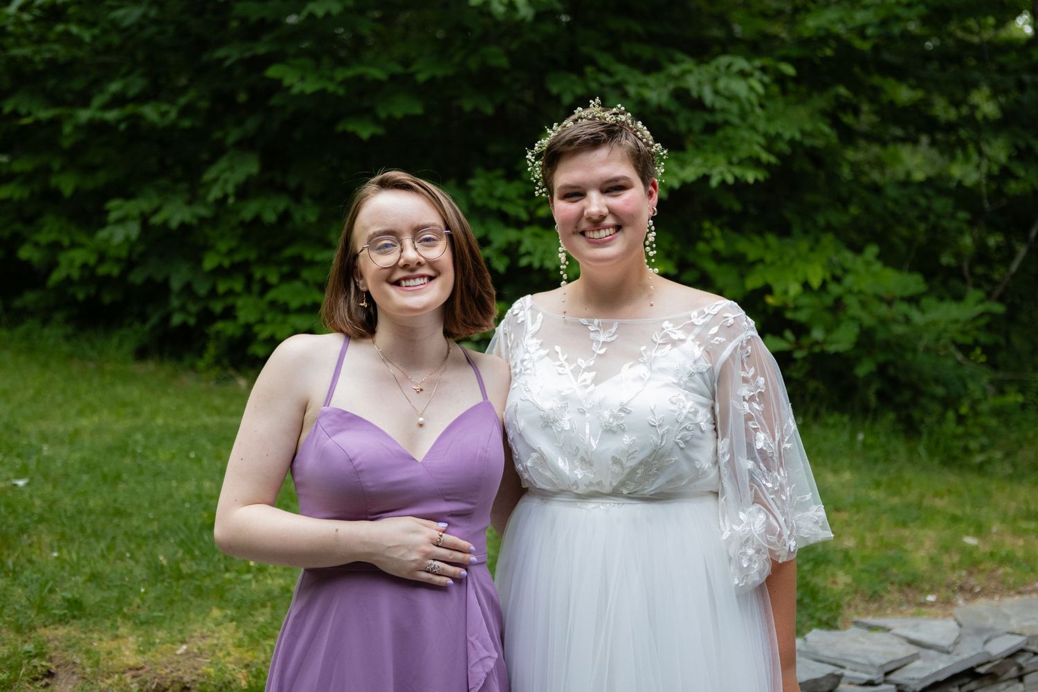 Outdoor wedding portrait with bride and attendant in purple dress against natural greenery.