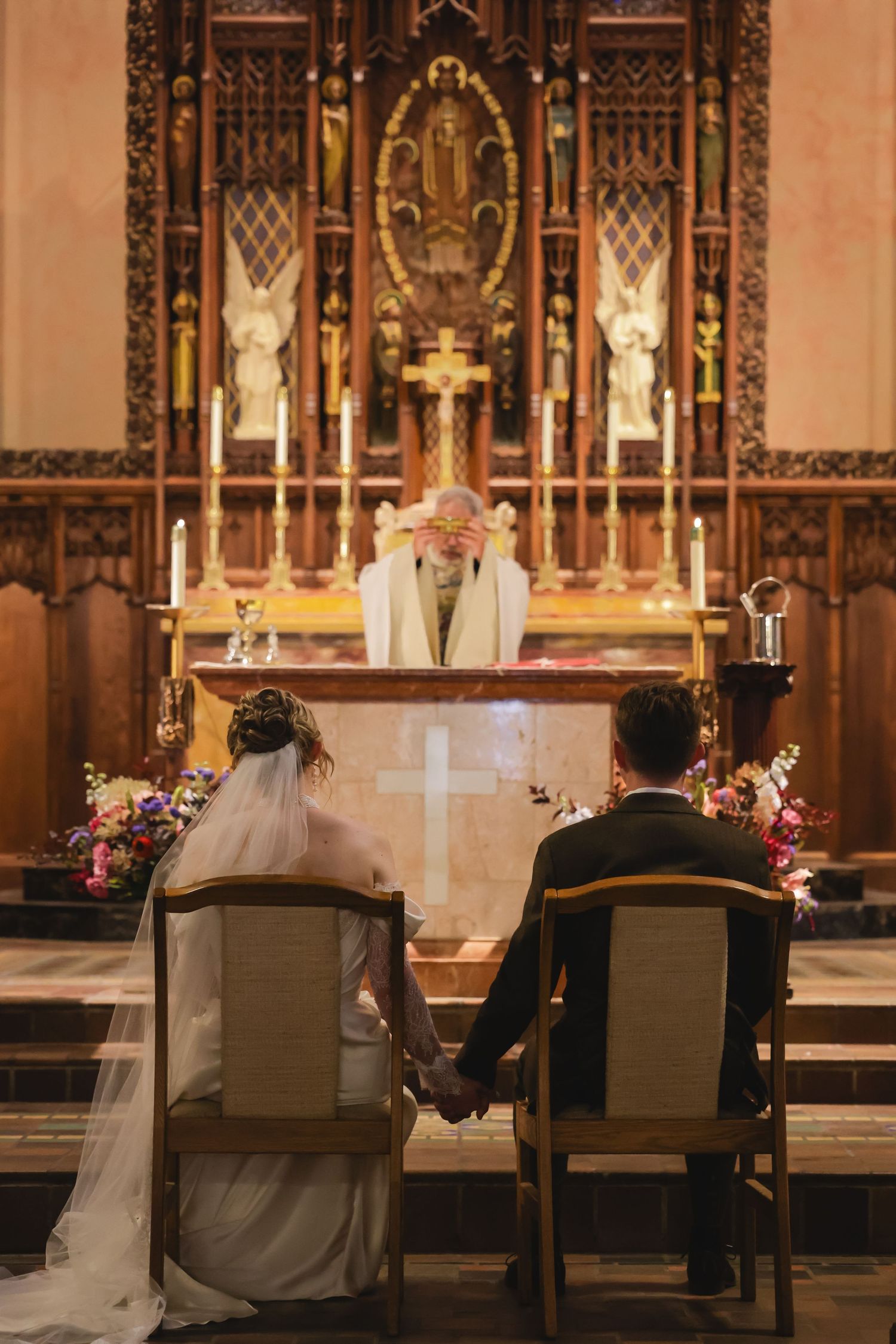 A couple holds hands during their wedding ceremony in an ornate cathedral with gothic architecture and lit candles at the altar.