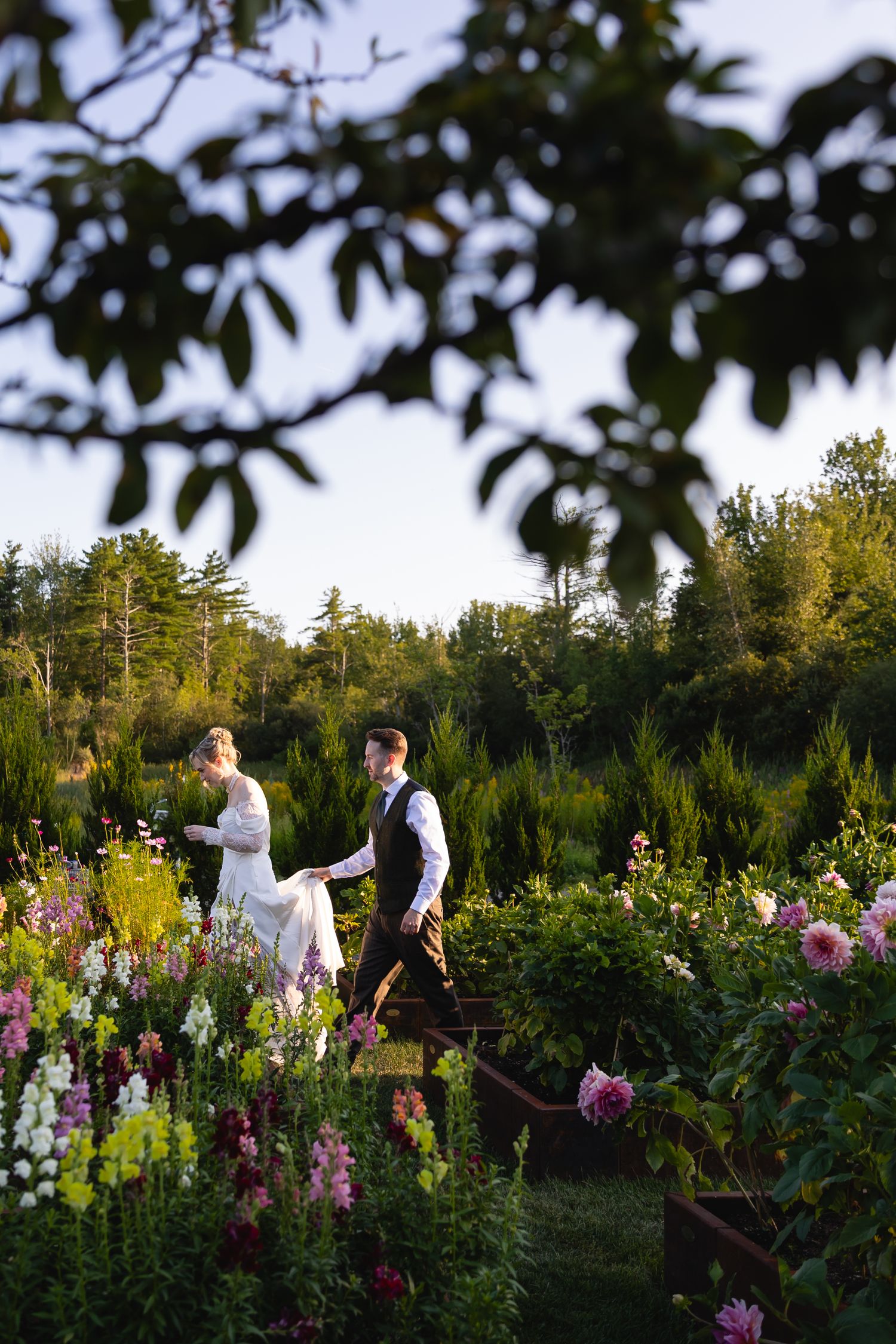 Two people stroll through a lush garden filled with pink and purple flowers at sunset, surrounded by tall trees.