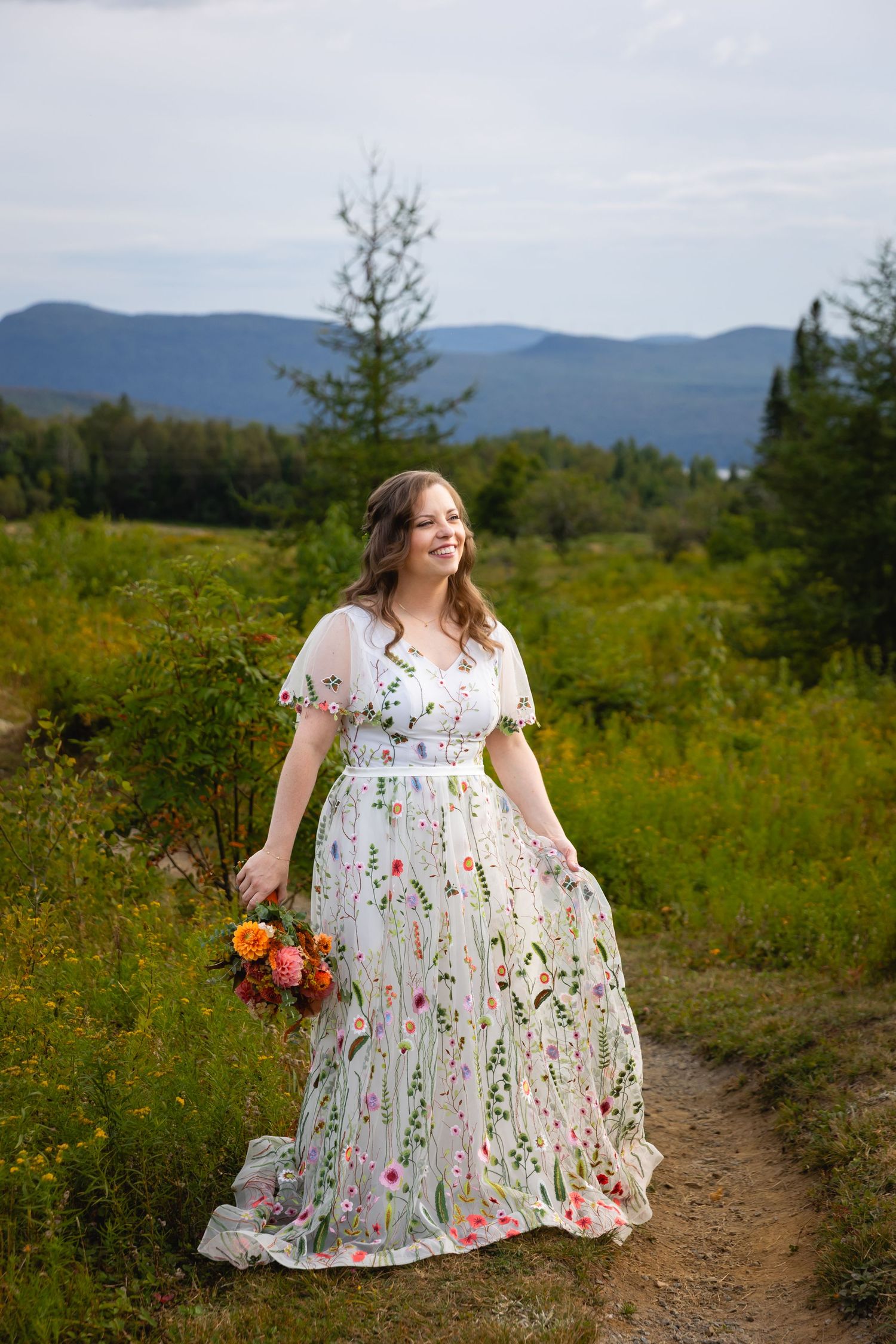 A floral embroidered white dress flows gracefully on a mountain trail surrounded by lush greenery and distant peaks.