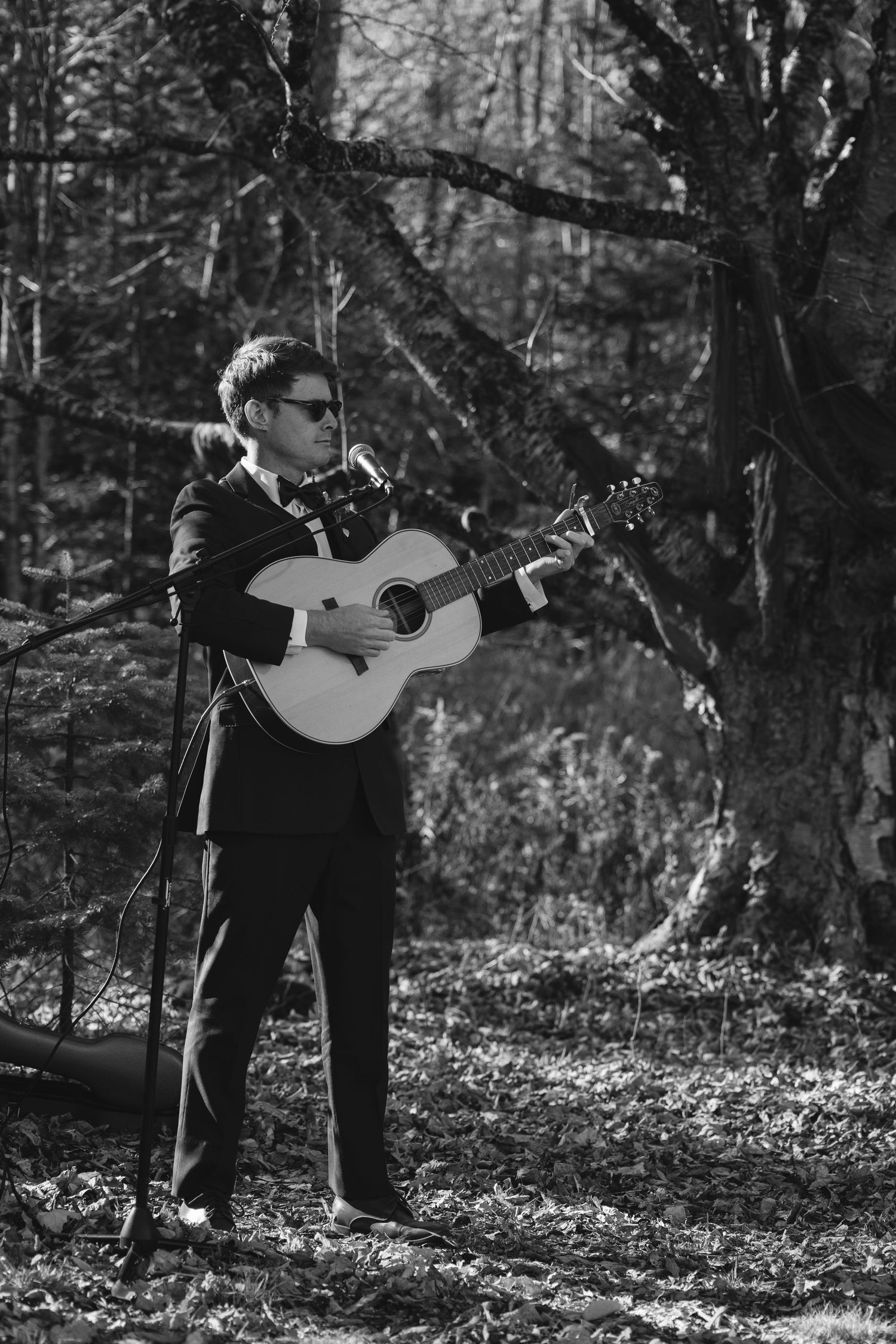 A musician in dark attire plays an acoustic guitar while standing in a wooded forest setting in black and white.