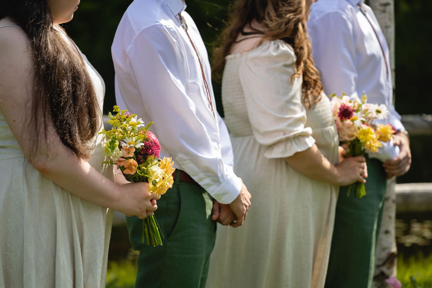 Wedding party members in white and green attire stand in line holding yellow bouquets during outdoor ceremony.