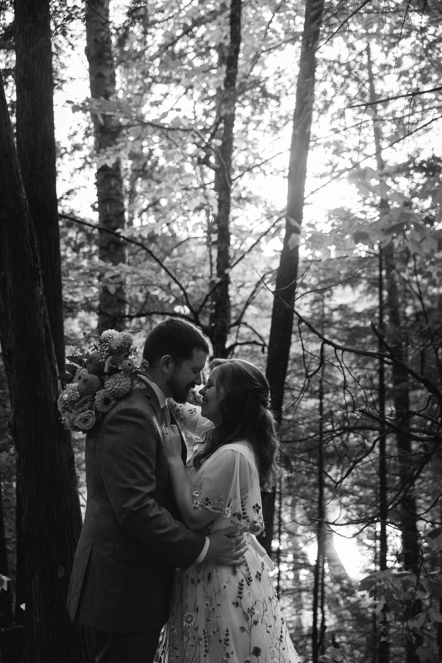 A romantic black and white forest scene with a couple embracing among tall trees and dappled sunlight.