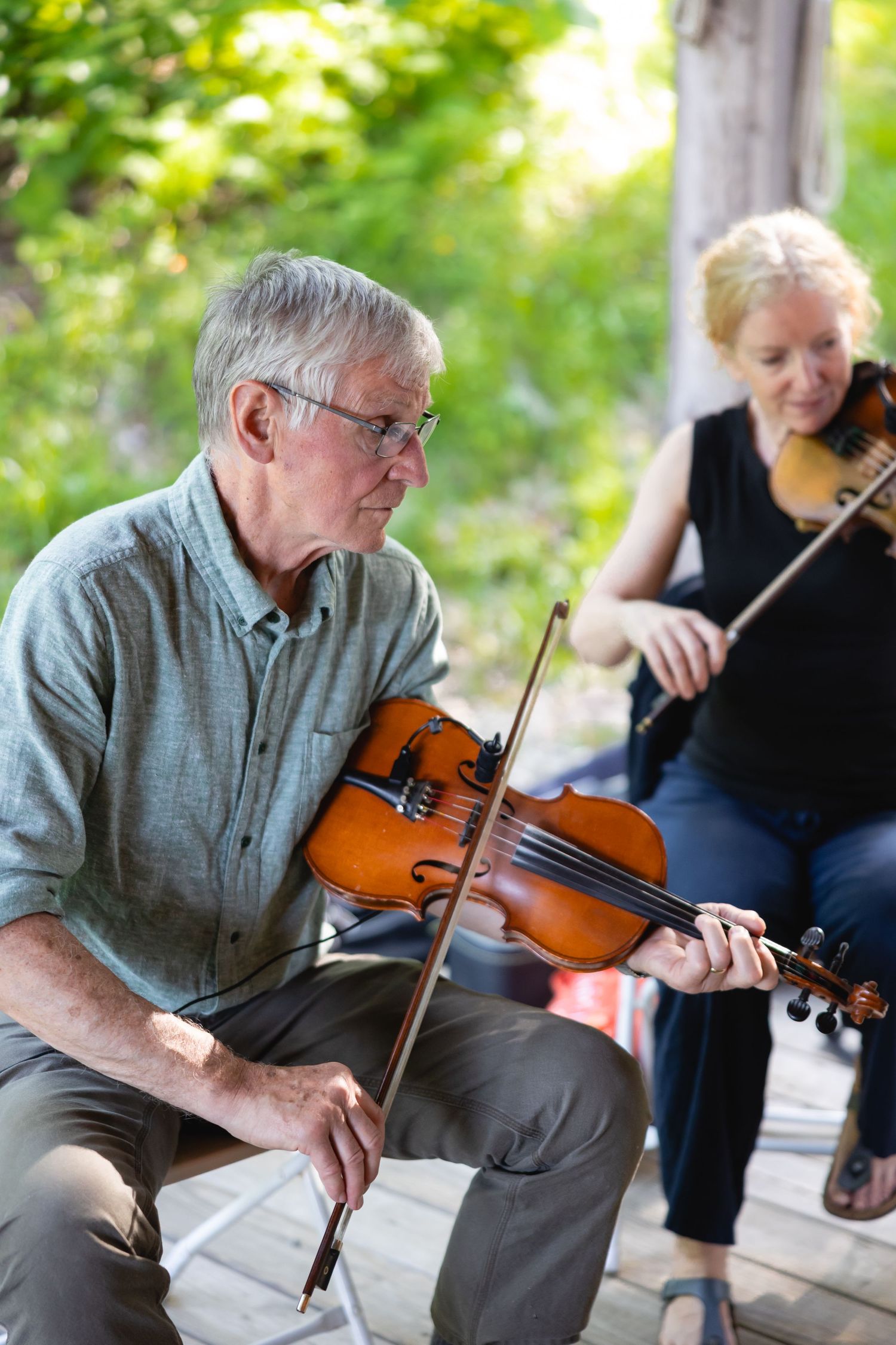 Two musicians playing violins together on a porch in a casual outdoor setting.