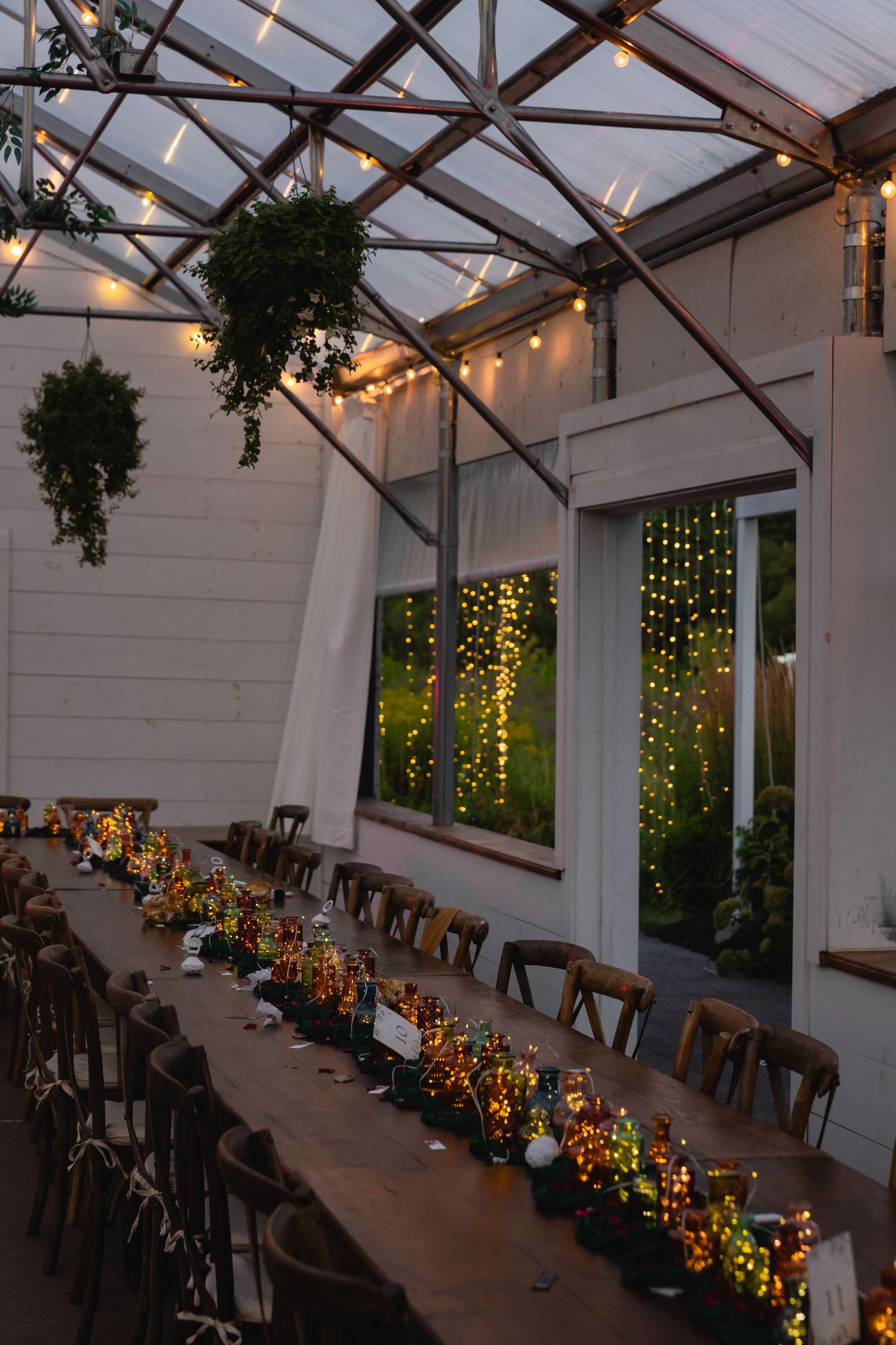 Long banquet tables set in a greenhouse venue with twinkling lights and hanging plants create an enchanting dining atmosphere.