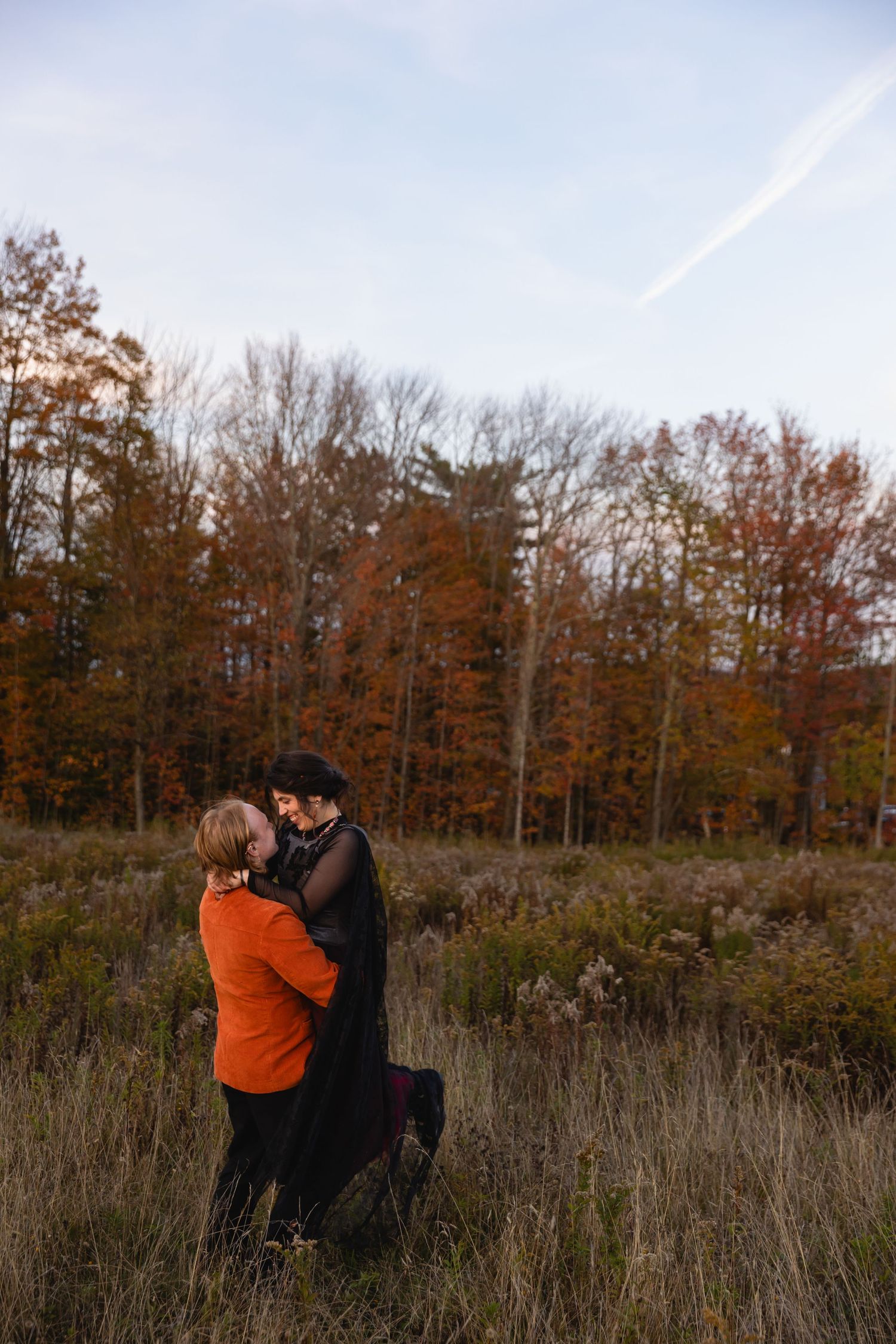 A couple embraces in a serene autumn field with tall grass and rustic orange trees in the background.