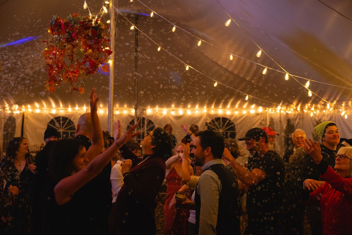Crowd of party guests dancing under a white tent illuminated by warm string lights and festive decorations.