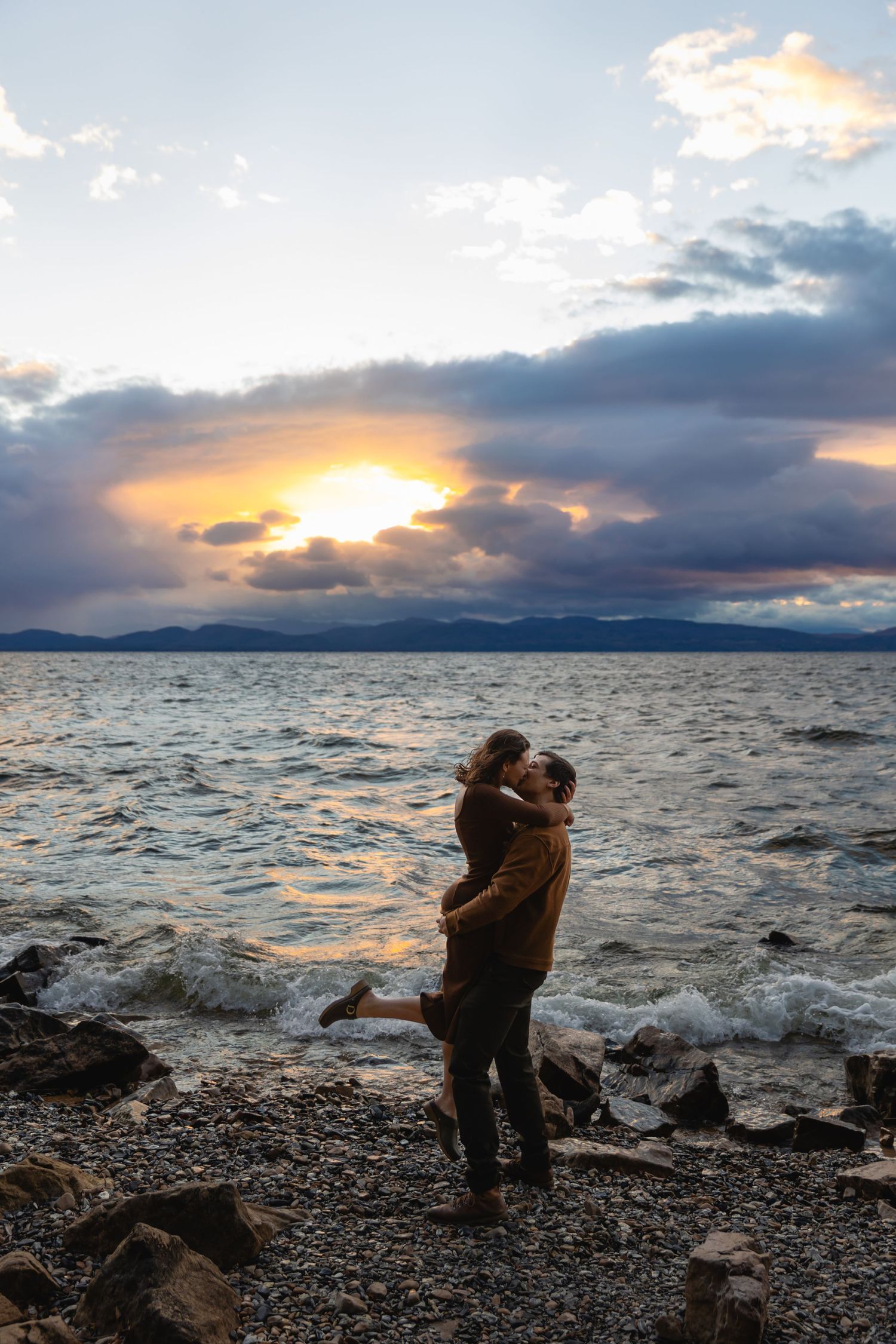 Couple shares a romantic embrace on a rocky shoreline during a dramatic colorful sunset.
