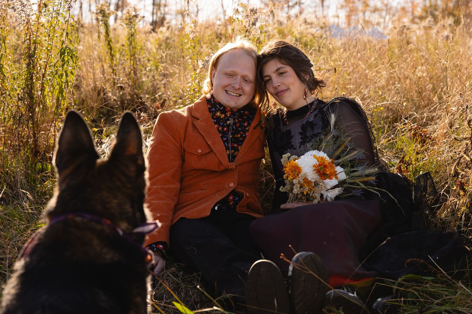 Happy couple sits together in autumn field with German Shepherd dog and bouquet of white flowers.