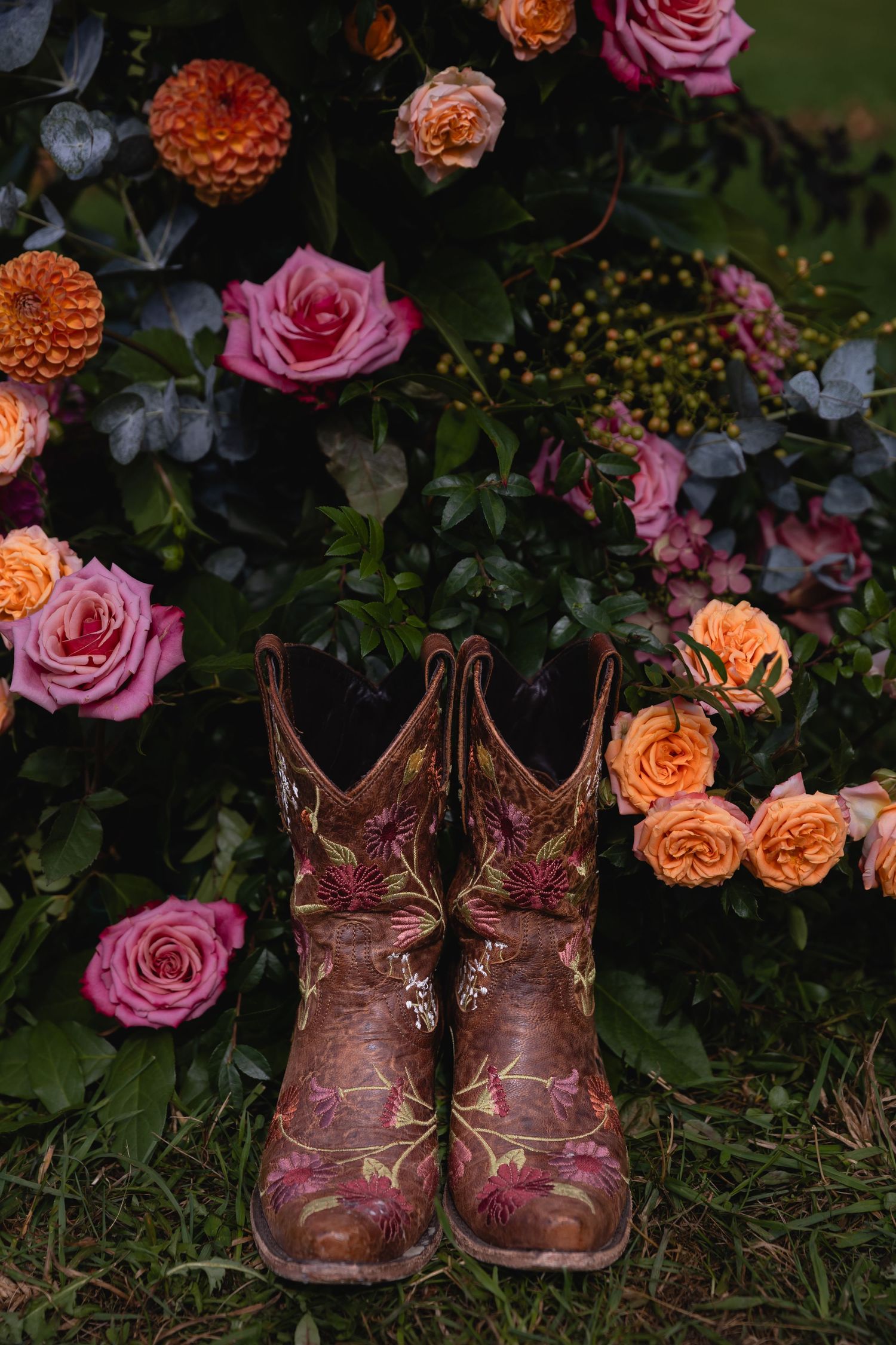 Embroidered cowboy boots surrounded by vibrant pink and orange roses in a lush floral arrangement.