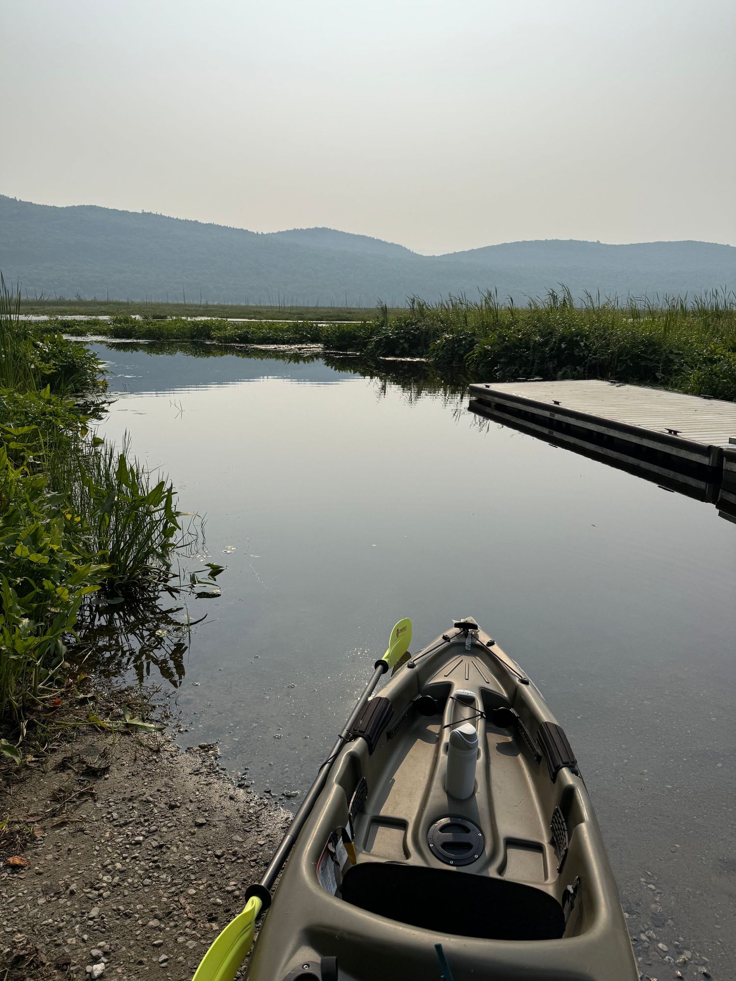 Kayak resting on a rocky shoreline with calm water, marsh grass, and misty mountains in background.