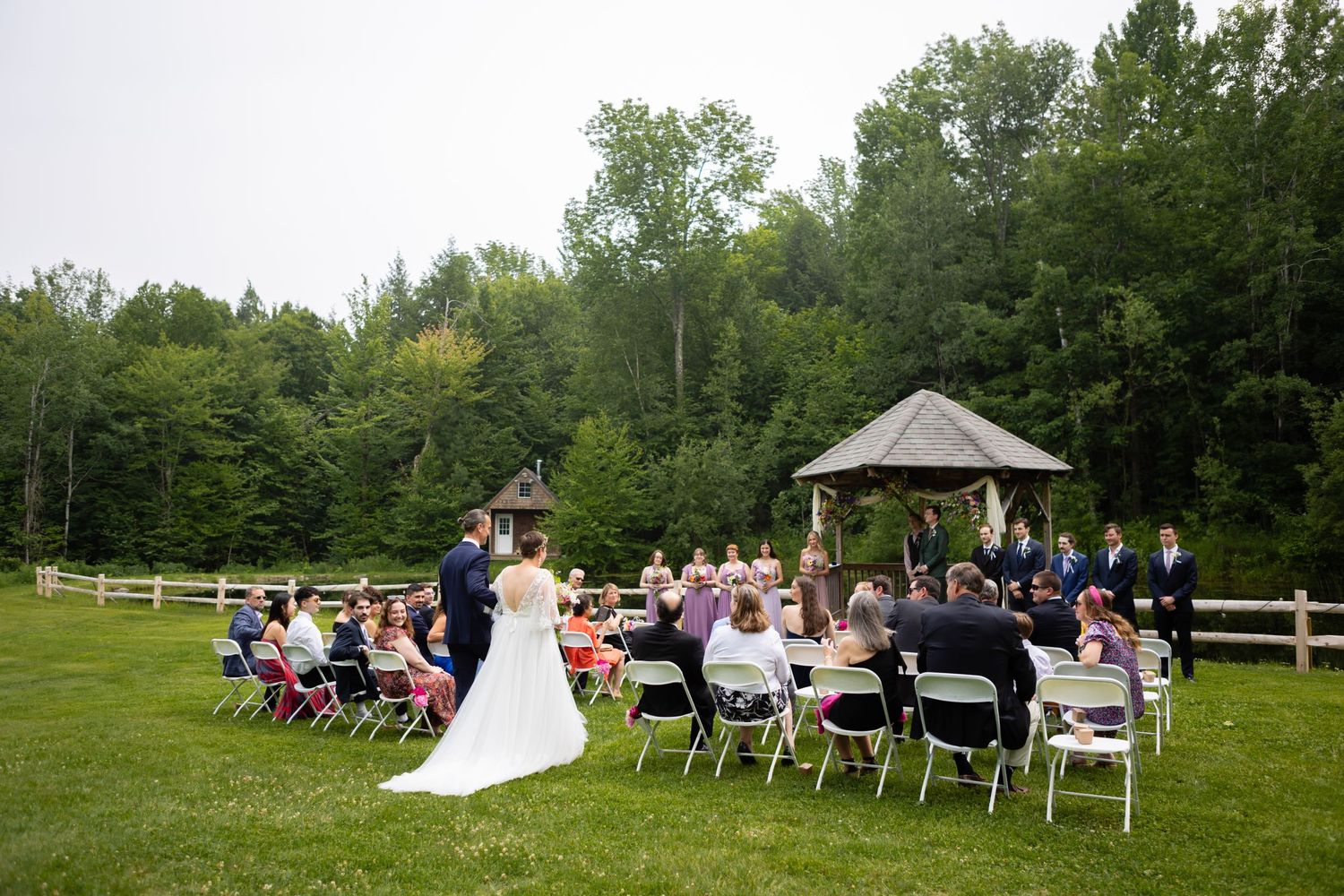 Wedding ceremony setup with white chairs and gazebo in a scenic outdoor forest setting.
