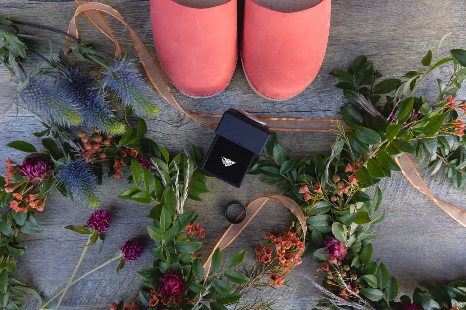 Pink slippers, ring box and floral arrangements laid out on rustic wooden floor.