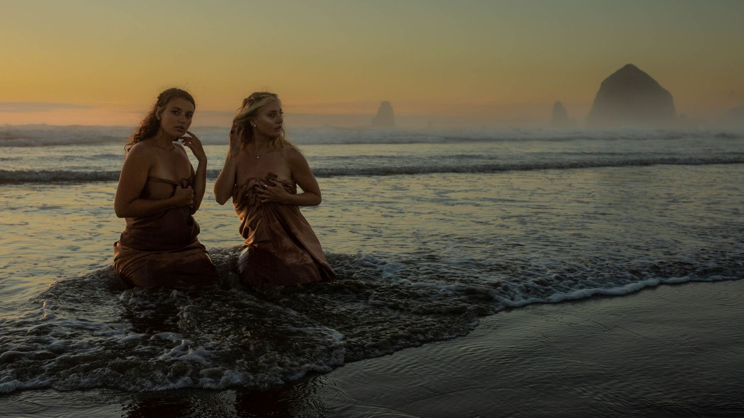 Two figures sit in flowing dresses at the ocean's edge during golden hour with Haystack Rock in the distance.