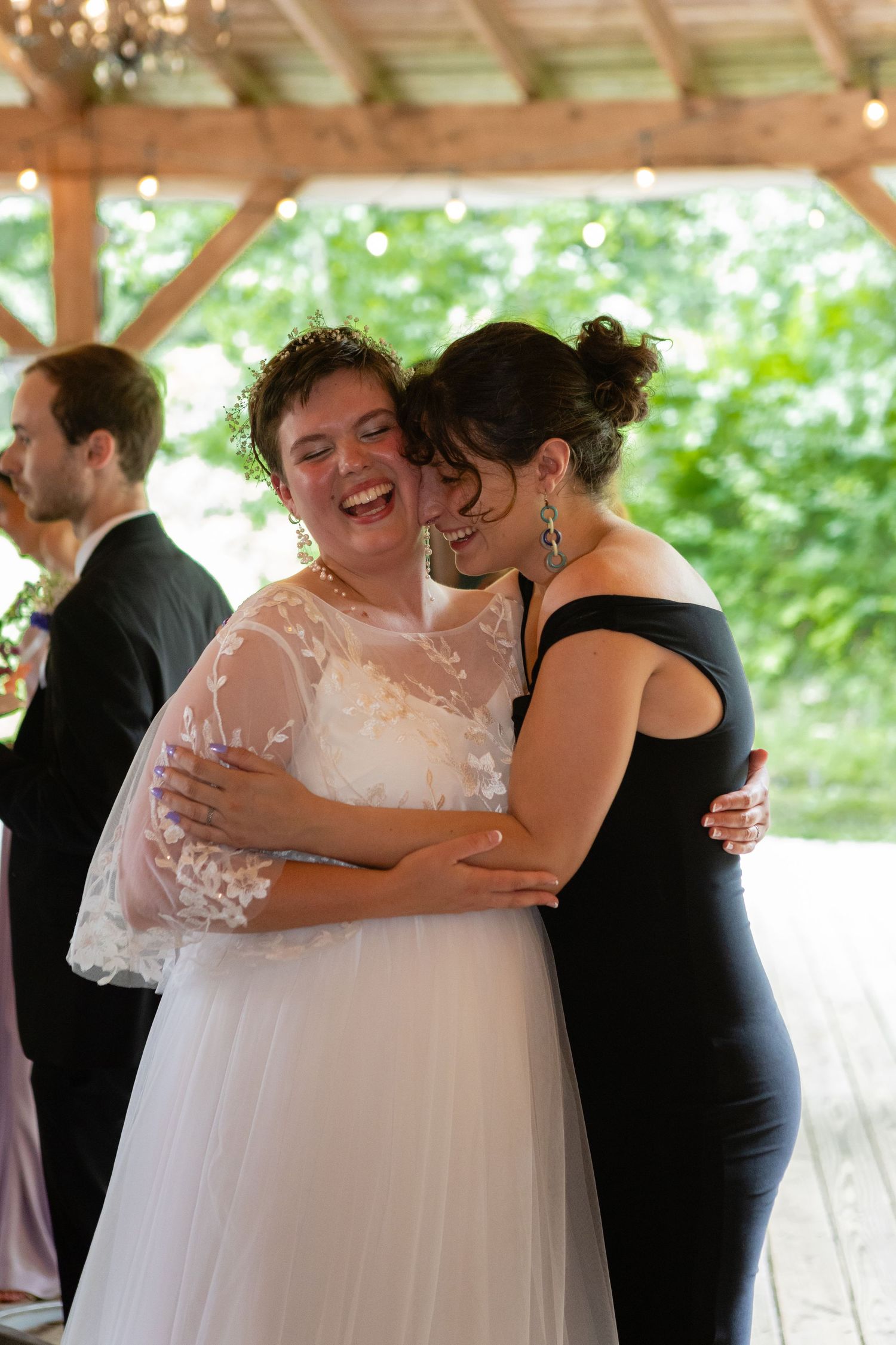 Wedding celebration with bride in white lace dress embracing guest in black dress under covered patio.