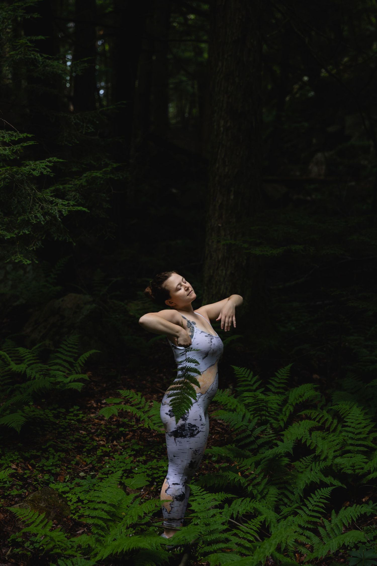 Woman in floral jumpsuit poses with arms crossed in a dark green fern-filled forest.