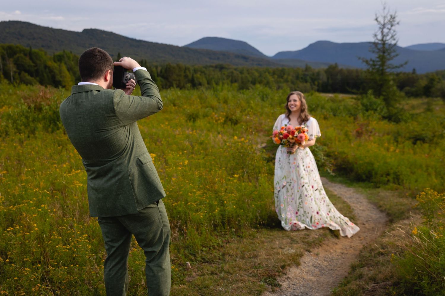 A couple in wedding attire poses together in a scenic mountain meadow filled with wildflowers at sunset.