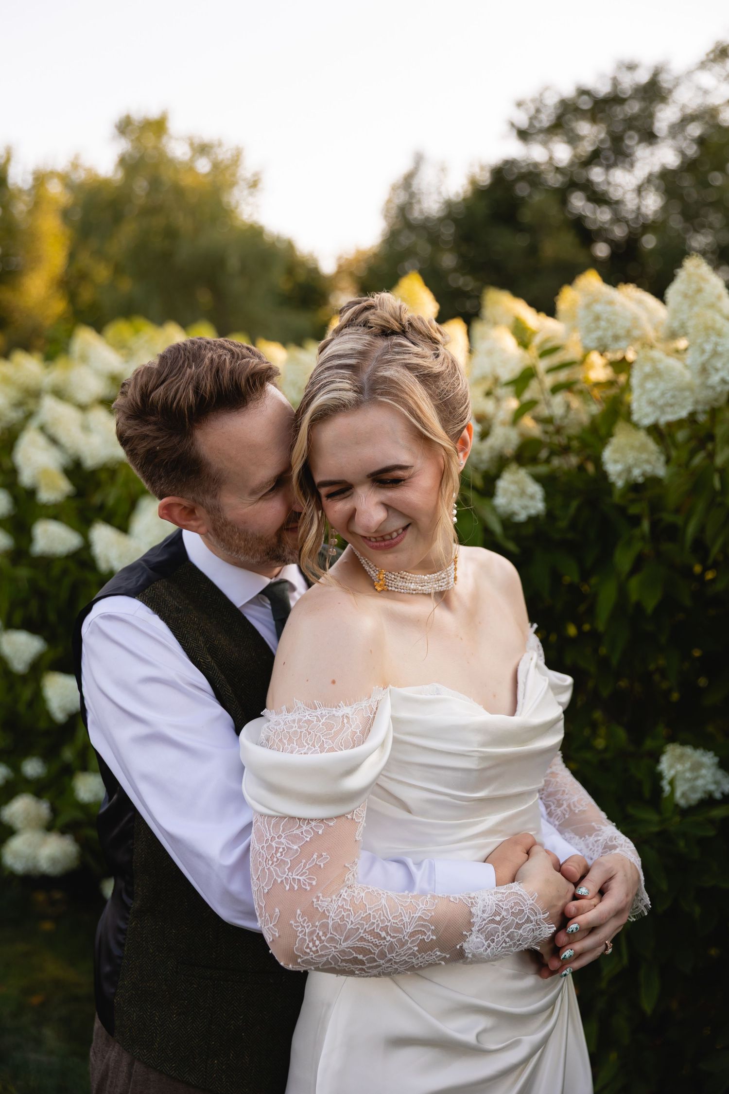 A romantic wedding couple embraces outdoors against a backdrop of white hydrangea bushes at sunset.