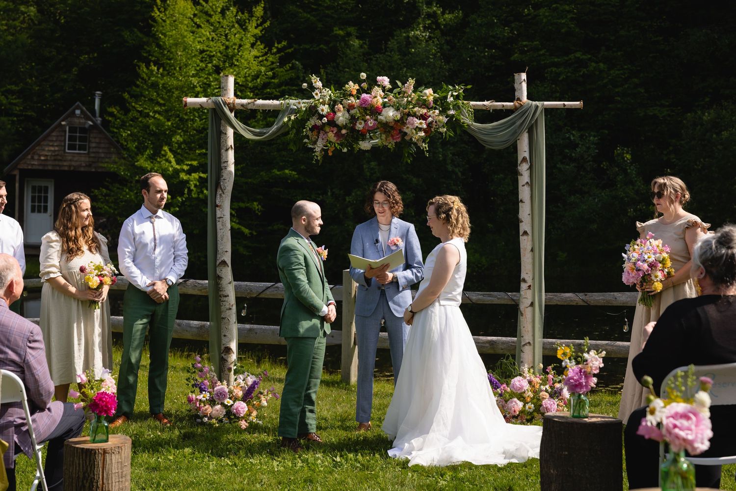 Wedding ceremony takes place outdoors with wooden cross altar decorated in pink and white flowers.