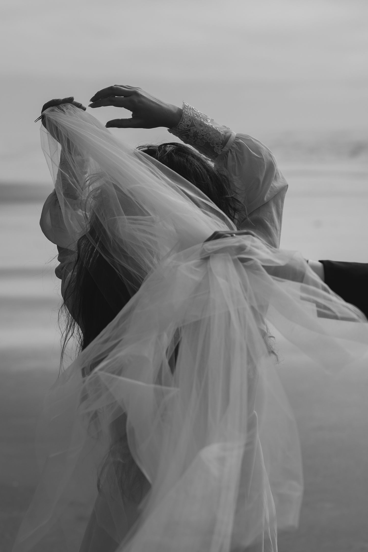 A figure draped in flowing white tulle fabric moves gracefully against a moody gray sky in black and white photography.