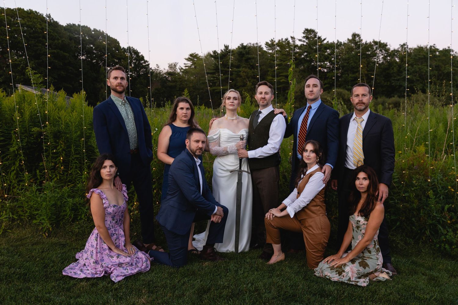 A wedding party poses together outdoors at dusk with a lush green field and treeline in the background.