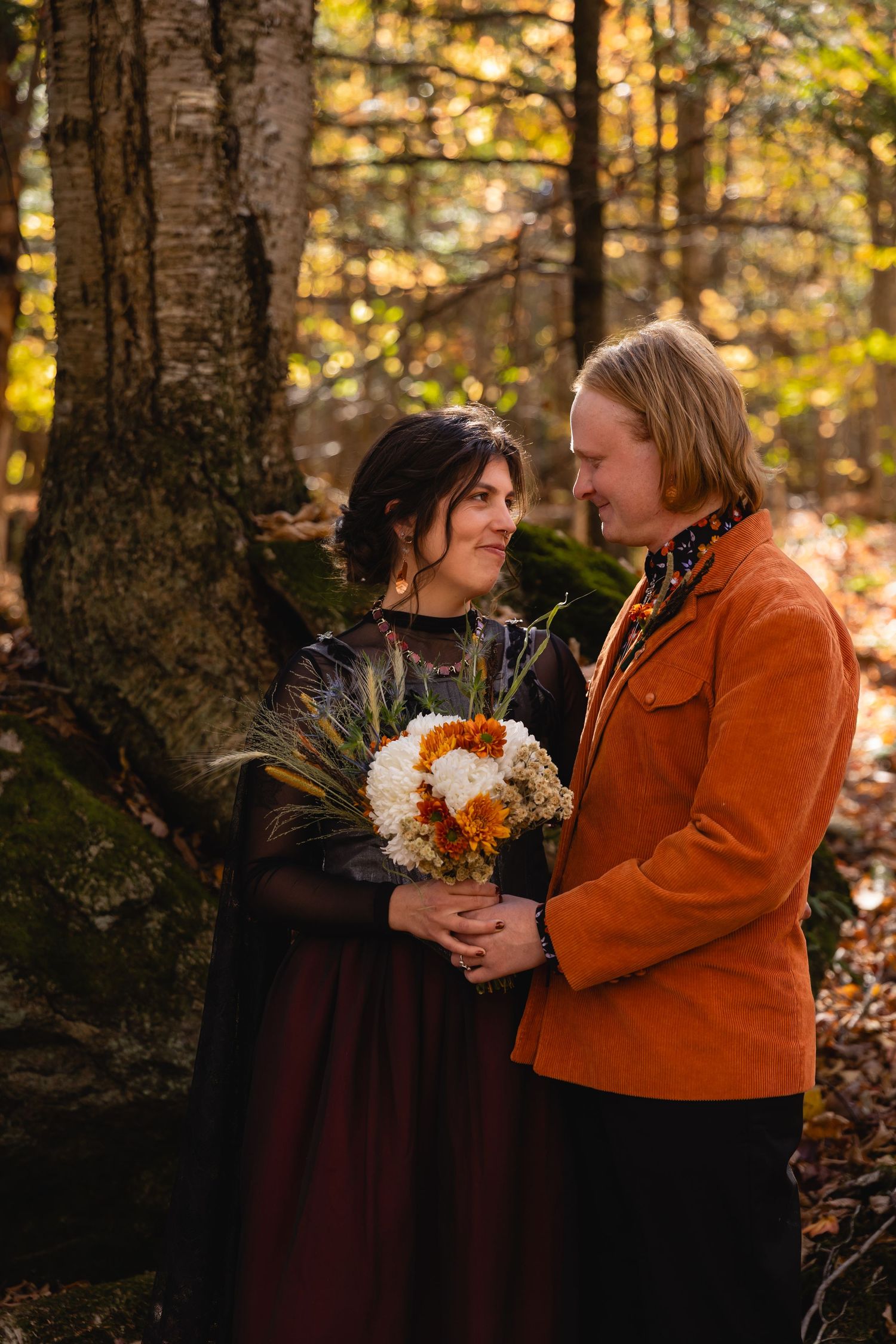 A couple shares an intimate autumn forest moment while holding a rustic bouquet of white flowers and dried grasses.
